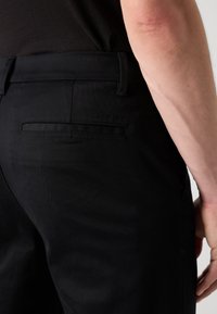 Zwarte broek met een gladde textuur, voorzien van een achterzak en subtiele merkvermelding bij de tailleband. Perfecte pasvorm met een strakke uitstraling.