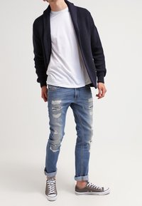 Cárdigan de punto azul marino, camiseta blanca, vaqueros azul claro desgastados y zapatillas grises. El cárdigan presenta un cuello de chal y puños acanalados.