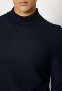 Tummansininen neulepaita, jossa on korkeakauluksellinen muotoilu ja ribbireunat kauluksessa sekä olkasaumoissa. Rinnassa on hillitty logopainatus.