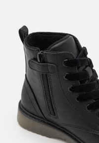 Černá kožená kotníková bota s měkkou vnitřní podšívkou, plochou podrážkou, šněrováním a bočním zipem pro snadné obouvání. Hladký povrch s detaily šití.