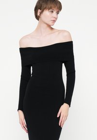 Rochie neagră, cu umerii la vedere, cu mâneci lungi și strâmte, realizată dintr-un material elastic; textură netedă și siluetă aderentă.