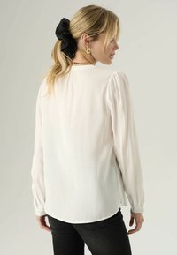 Bílá halenka s dlouhými rukávy, hladkým povrchem, jemným leskem, knoflíkovými manžetami a mírně zaobleným lemem, doplněná černou gumičkou do vlasů.