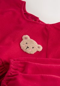 Vestito rosso in velluto a coste con un'applicazione di un orsetto di peluche. L'orsetto ha occhi cuciti e un naso rosa, posizionato vicino allo scollo.