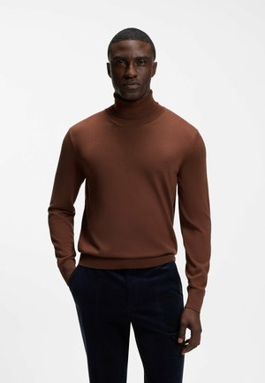 Homme portant un pull à col roulé marron et un pantalon foncé, debout avec une main dans la poche devant un fond blanc uni.