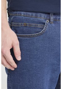 Temno modri kavbojki iz jeansa z gladko teksturo, z sprednjim žepom in vidnimi šivi v kontrastni barvi. Roka počiva na tkanini.