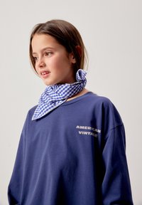 Girl wearing a blue "American Vintage" sweatshirt and a blue-and-white checkered neck scarf, looking to the left against a plain background.