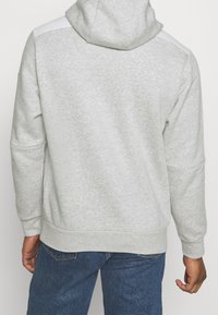 Gray hoodie with a textured finish, featuring a white panel on the shoulders and ribbed cuffs. Visible seam details and relaxed fit.