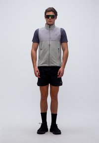 Moški stoji oblečen v sivo brezrokavnik z zadrgo čez mornarsko modro srajco, črne kratke hlače, črne nogavice, črne superge in odsevna športna sončna očala.