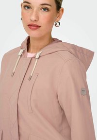 Jeune femme à la peau claire et aux yeux bleus portant des boucles d'oreilles argentées et une veste à capuche rose pâle avec des cordons de serrage et un petit logo sur la manche.