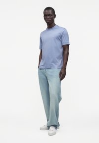 Hombre con camiseta azul claro de manga corta, vaqueros holgados de lavado claro y zapatillas blancas, de pie frente a un fondo blanco liso.