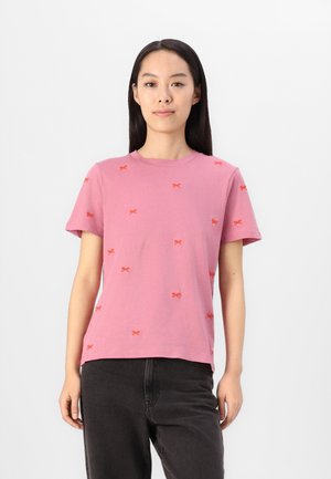 T-shirt en coton rose à manches courtes, avec de petits motifs de nœuds rouges sur toute la surface. Encolure ronde et coupe décontractée. Pantalons noirs portés.