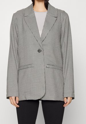 Frau trägt einen schwarz-weißen Hahnentritt-Blazer mit einem einzelnen schwarzen Knopf über einem hellgrauen Oberteil und schwarzen Hosen.