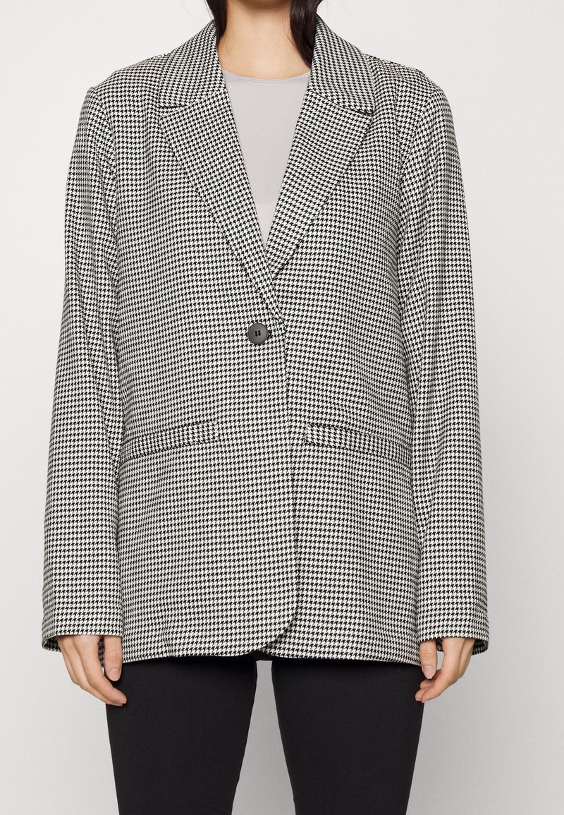 Femme portant un blazer pied-de-poule noir et blanc avec un seul bouton noir sur un haut gris clair et un pantalon noir.