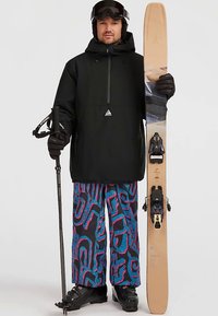 Man wearing black jacket, colorful patterned ski pants, helmet, gloves, holding ski pole and beige alpine ski.