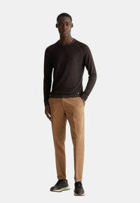 Maglione lungo marrone a maniche lunghe con girocollo, abbinato a pantaloni beige e sneakers nere. Tessuto liscio; design slim-fit con dettagli minimi.