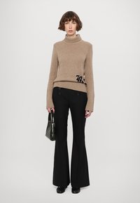 Beige gerippter Rollkragenpullover mit schwarzer Stickerei, kombiniert mit schwarzen Schlaghosen. Das Modell hält eine dunkelgrüne Handtasche. Glatte Texturen.