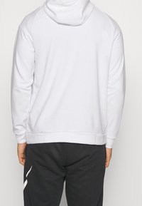 Personne portant un sweat à capuche blanc uni et un pantalon noir avec un graphisme blanc sur la jambe gauche, vue de dos.