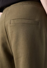 Gros plan d'un pantalon vert olive montrant une poche passepoilée arrière et une partie d'un haut noir porté par une personne.