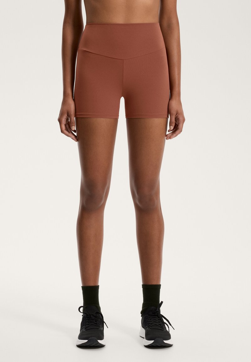 Högmidjade, rostfärgade träningsshorts med stretchy tyg och en slät textur; parat med svarta sneakers och gröna strumpor.