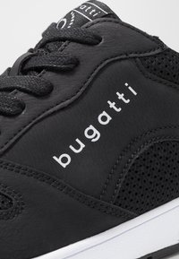 Black athletic shoe featuring a smooth leather upper, mesh accents, and white logo text. Equipped with black laces and a white rubber outsole.