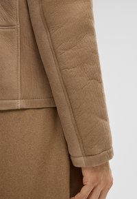 Manica di giacca marrone dalla texture morbida con cuciture visibili e polsino ripiegato. Il tessuto sembra leggero e leggermente stropicciato.