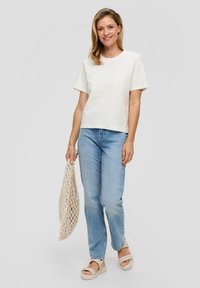 Camisa blanca de manga corta con rayas amarillas claras, corte relajado, combinada con unos jeans de mezclilla azules y sandalias trenzadas beiges, sujetando una bolsa de mimbre.