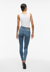 Calças de ganga azuis de cintura alta com uma textura suave, bolsos traseiros e um rótulo de pele castanha na cintura. Usadas com uma t-shirt branca sem mangas.