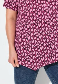 Roze en donkerpaarse top met een hartjespatroon in verschillende tinten roze, vergezeld van blauwe denim jeans en zichtbare stikdetails.