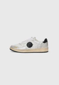Zapatilla blanca y plateada con parte superior texturizada, logo circular negro, letras doradas y suela de goma. Los cordones pasan por la lengüeta.