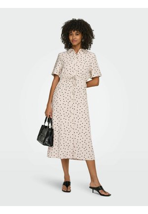 Nainen, jolla on kiharat hiukset ja joka käyttää beigeä, lyhythihaista, napitettua pilkullista mekkoa. Hän pitää käsissään mustaa käsilaukkua ja käyttää mustia korkokenkiä.