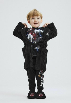 Schwarzer Fleece-Mantel mit Taschen, über einem Grafik-T-Shirt mit einem Superhelden getragen. Schwarze Hosen mit grafischem Druck, flauschige schwarze Hausschuhe.