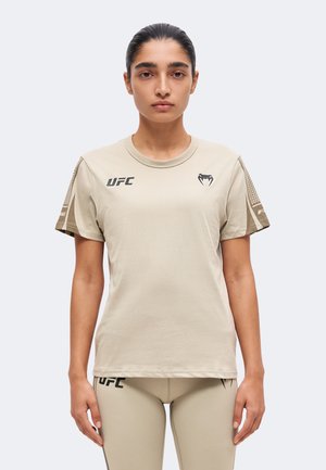 Жена с тъмна прическа на опашка, облечена в бежова спортна тениска и клин на UFC, стои изправена с лице към камерата на прост фон.