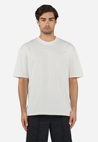 Tricou alb din bumbac, cu o croială lejeră, mâneci scurte, decolteu rotund și un logo brodat subtil pe piept. Textură netedă.