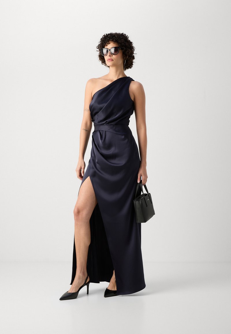 Granatowa satynowa gown z jednym ramieniem, z wysokim rozcięciem, z przylegającą talią, zestawiona z czarnymi szpiczastymi szpilkami i małą czarną torebką.