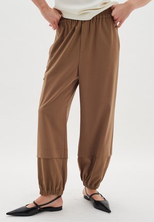 Pantalons marron texturés à taille élastique et poignets froncés, coupe large; associés à des ballerines noires à bouts pointus.
