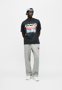 Μαύρο γραφικό μπλουζάκι με εκτύπωση σύννεφου και έντονο κείμενο, συνδυασμένο με ανοιχτό γκρι φόρμα και λευκά αθλητικά παπούτσια. Μπλε καπέλο φορεμένο ανάποδα.