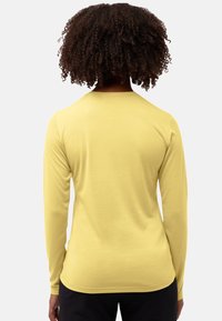 Person mit lockigem Haar, der ein einfaches, gelbes Langarmshirt und schwarze Hosen trägt, steht mit dem Rücken zu einem weißen Hintergrund.