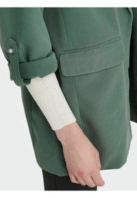 Vihreä, teksturoiduinen takki, jossa on käärityt hihat ja etutasku. Valkoinen ribbikuvioinen villasukka näkyy sen alla. Klassinen muotoilu, ilman koristeluja.