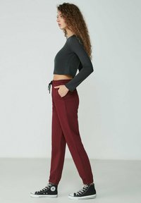 Schwarzes, kurzes Langarmoberteil kombiniert mit burgunderfarbenen Jogginghosen. Die Hose hat einen elastischen Bund und Bündchen. Schwarze Sneaker runden den Look ab.