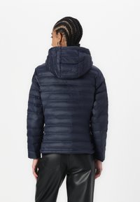 Marine blauwe pufferjack met capuchon, voorzien van een gestikte textuur, lange mouwen en een getailleerd silhouet. Gecombineerd met zwarte neplederen broeken.