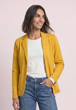 Kvinde med skulderlangt brunt hår iført en gul blazer, hvid skjorte og blå jeans, smilende og kigger til siden med den ene hånd i lommen.