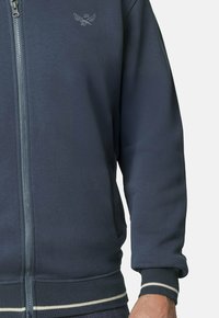 Sudadera con cremallera en color azul marino, con puños y dobladillo acanalados, que presenta un logotipo sutil en el pecho y franjas blancas contrastantes en la cintura.