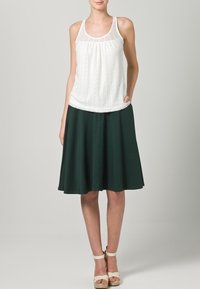 Top in pizzo bianco con corpino plissettato, abbinato a una gonna verde scuro a lunghezza ginocchio. Presenta una texture liscia e un design semplice.