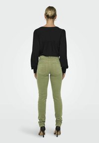 Blouse noire à manches longues avec un dos froncé, associée à un pantalon fitted vert clair et des chaussures à talons, vue de dos.