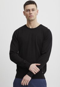 Svart genser med rund hals og lange ermer, med ribbestrikkede mansjetter og hem, laget av et mykt stoff. Har et lite logoakksent på den nederste siden.