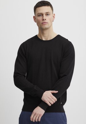 Svart genser med rund hals og lange ermer, med ribbestrikkede mansjetter og hem, laget av et mykt stoff. Har et lite logoakksent på den nederste siden.