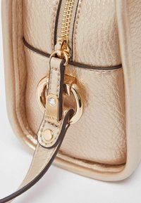 Primer plano de bolso metálico dorado que muestra material texturizado, cierre de cremallera, detalles de herrajes dorados y un accesorio decorativo en forma de anillo.