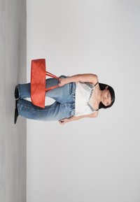 Sieviete stāv neformālā apģērbā ar baltu bezpiedurkņu augšu, vaļīgām zilām biksēm, melnām kurpēm, tur rokā lielu oranžu somu uz vienkārša pelēka fona.