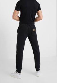 Svart bomullsblandning joggers med elastisk midja, sidofickor och guldlogotyp på bakfickan. Matchas med en åtsittande svart T-shirt.