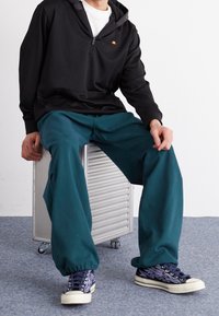 Felpa nera con zip, pantaloni baggy turchesi e sneakers navy con suole bianche. La felpa presenta un piccolo logo; i pantaloni hanno polsini elastici.